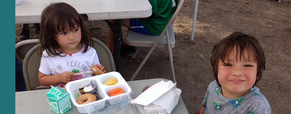 A young girl and her brother smiling and seated at a picnic table preparing to eat a box lunch
