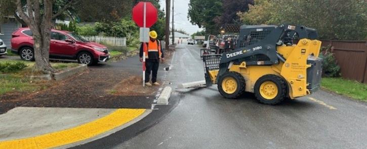 construction worker wearing a safety vest monitoring a small forklift at a curb cut