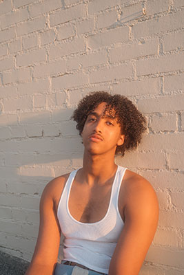 A young Black man leans against a wall at golden hour. He's wearing a white tank top and has long, curly hair.