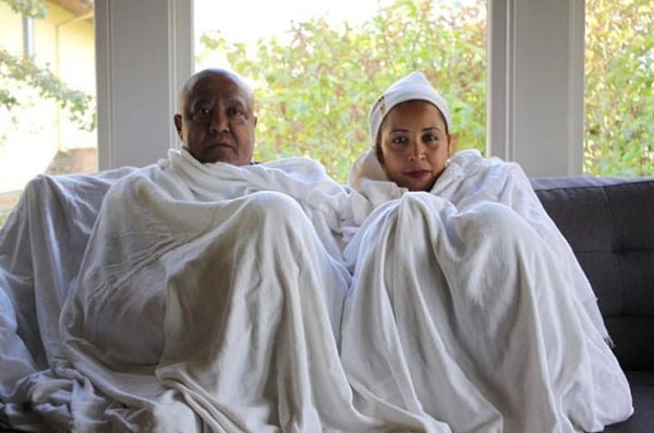 An Ethiopian couple wearing gabi sit on the couch looking into the camera.