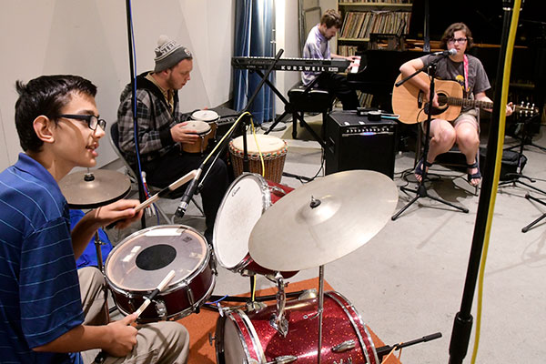Blind youth sit at a drum kit, bongos, piano, and guitar inside a studio.
