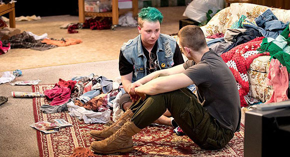 Two actors sit on the floor of the stage, one of them is trans