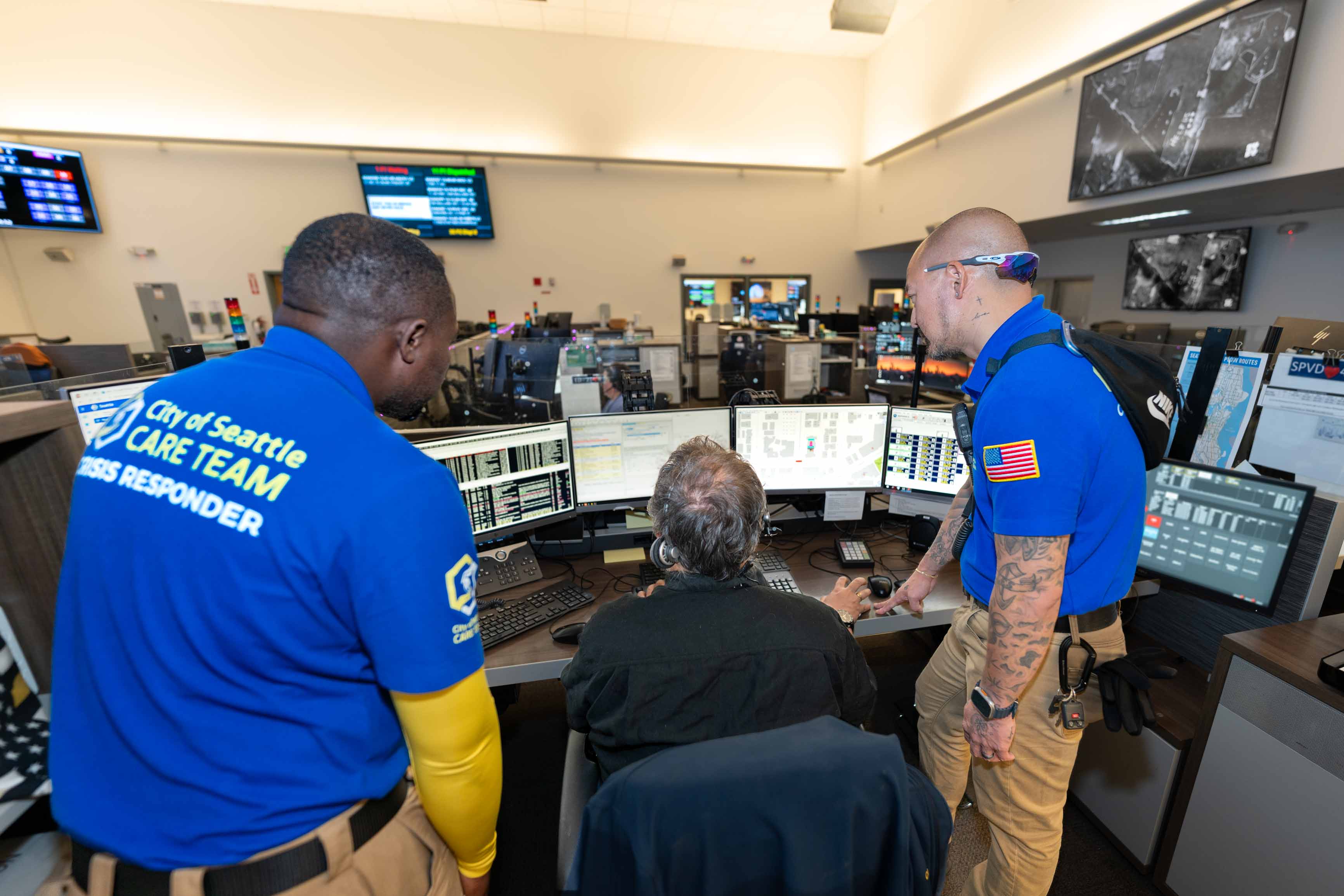 A 9-1-1 dispatcher and two CARE community crisis responders viewing multiple computer monitors on the desk of a Seattle 9-1-1 dispatcher