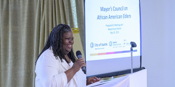 Smiling Black woman standing in front of a group of people and a screen with a slide showing Mayor's Council on African American Elders on it along with City of Seattle and Seattle human Services Department logos on it
