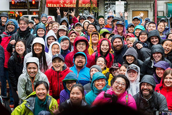 A large group of Asian, Pacific Islander, and Native Hawaiian people pose for a group shot in rain gear outside Mike's Noodles.