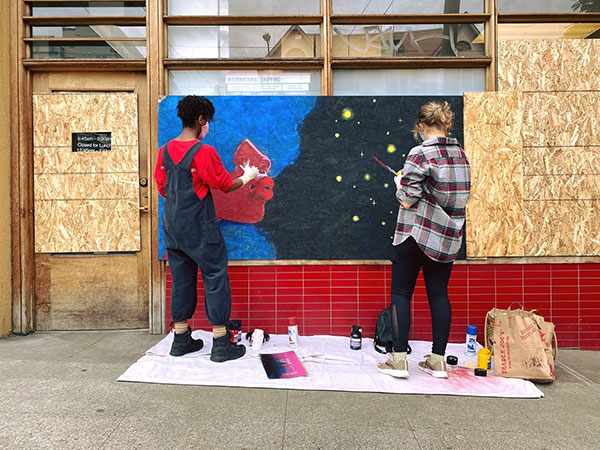 Two people paint a mural on a boarded up storefront.