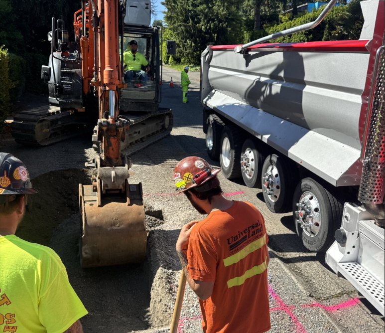 Construction workers and heavy equipment at an active work site near NE 104th St and 15th Ave NE. An excavator is digging into the roadway while crew members in safety gear monitor the process. A big truck is parked nearby.