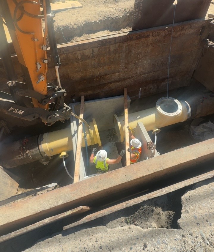 Construction workers install a large yellow water pipe underground, using heavy equipment to position and secure it inside a protective trench box. 