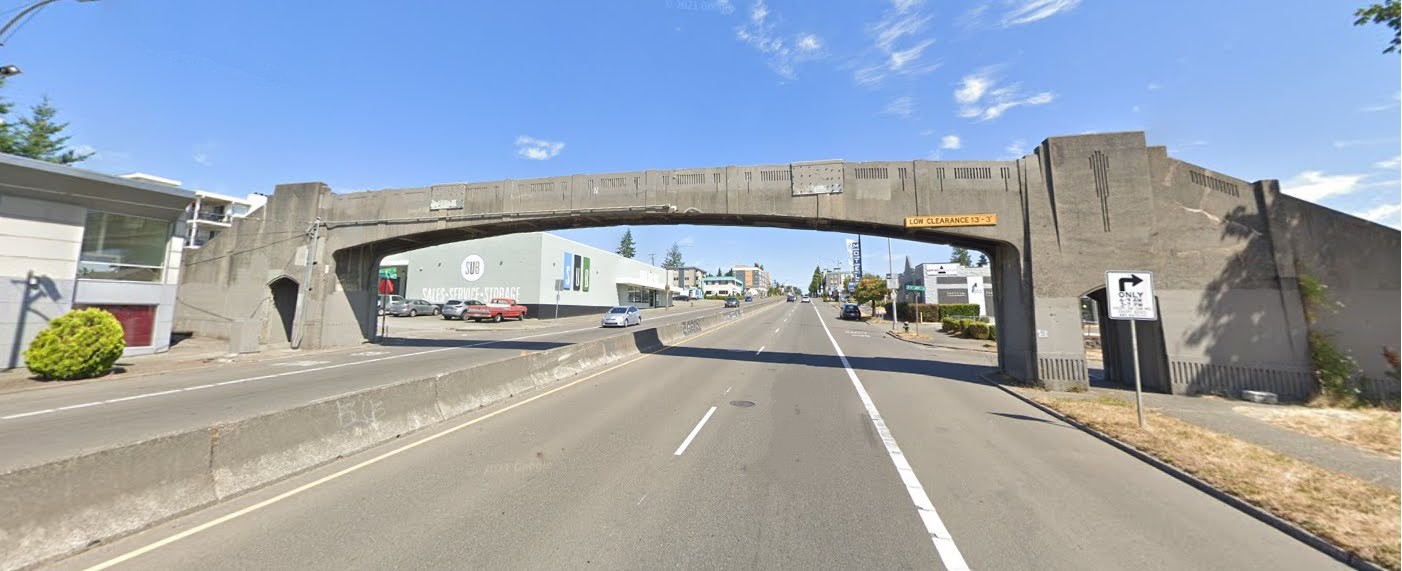 A concrete pedestrian bridge with an art deco design spans across a multi-lane road. The bridge shows visible wear, and a low clearance sign is posted