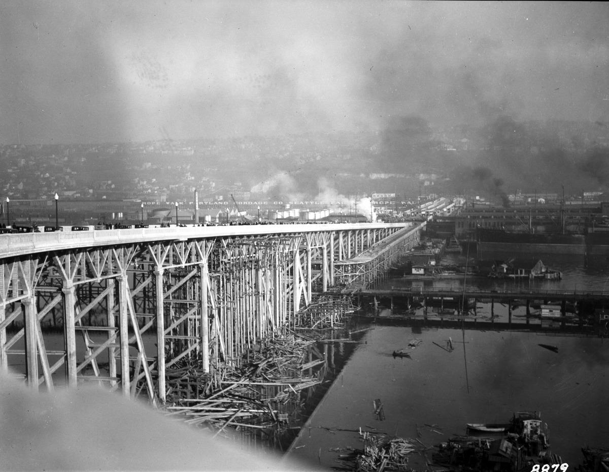 Historic image of the Magnolia Bridge spanning east over what is currently Terminal 91. Log piles are located at the base of the bridge, and there are smoky clouds in the sky.