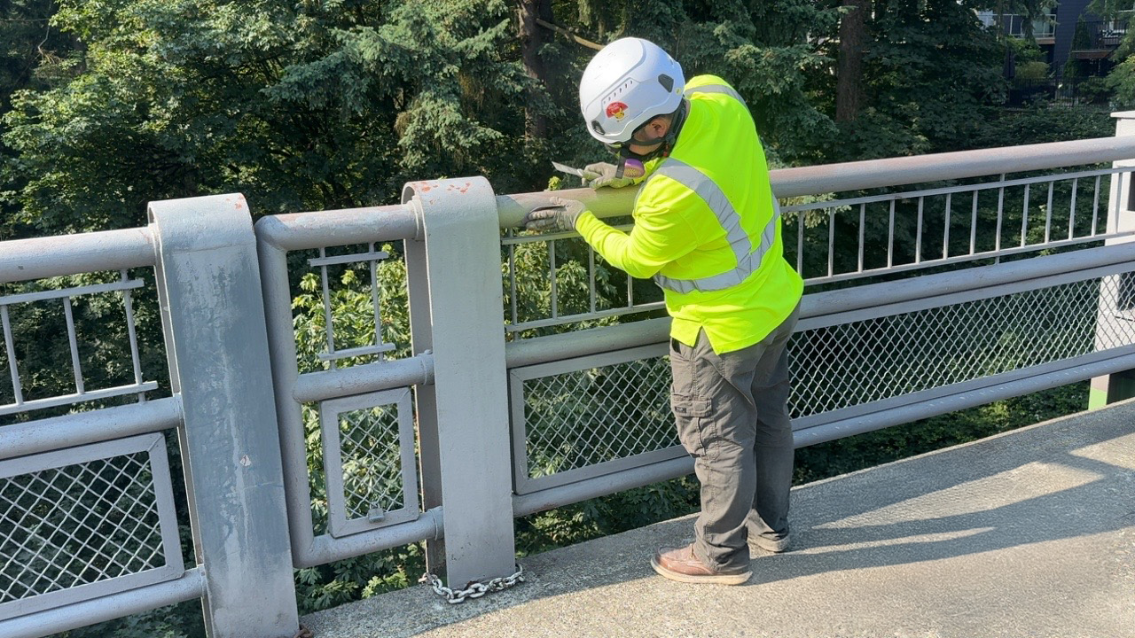 construction workers wearing safety gear sand and prepare the metal railing on a bridge surrounded by tall trees.