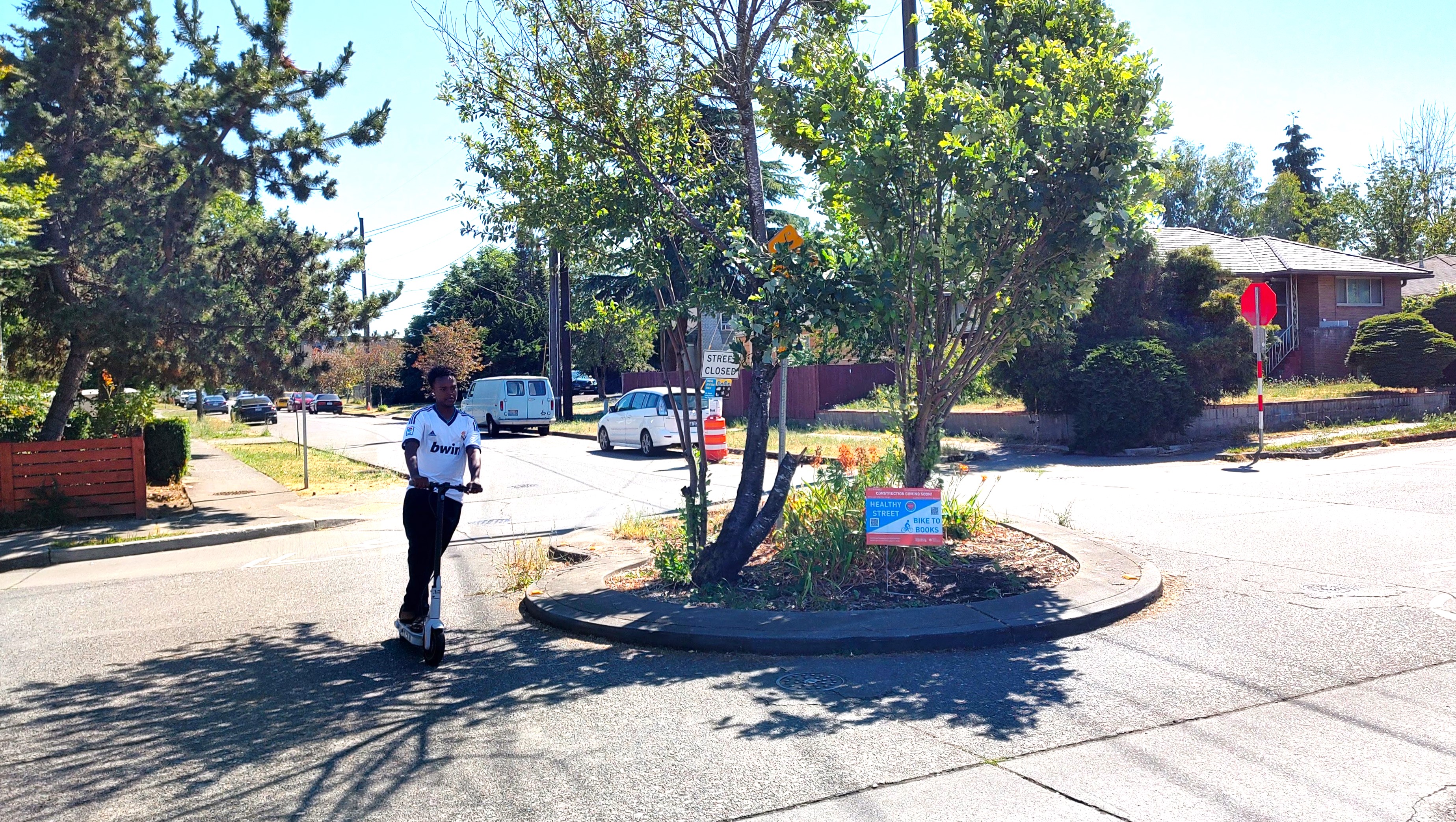 A young man on a scooter rolls past a traffic circle with a Healthy Street Coming Soon yard sign posted