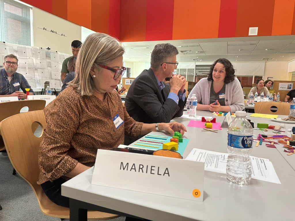 Three people are in the foreground, seated at a table in a school library. One is a Filipina woman in glasses and a brown shirt, looking at blocks and art supplies she is arranging on a piece of blue paper. The other two people are a white man and white woman, facing each other and talking. There are other people standing in the background.
