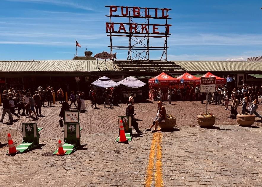 People walk through the market, behind planters with a sign in them that narrow the entry, and green barriers with cones on them block the rest of the entrance. 