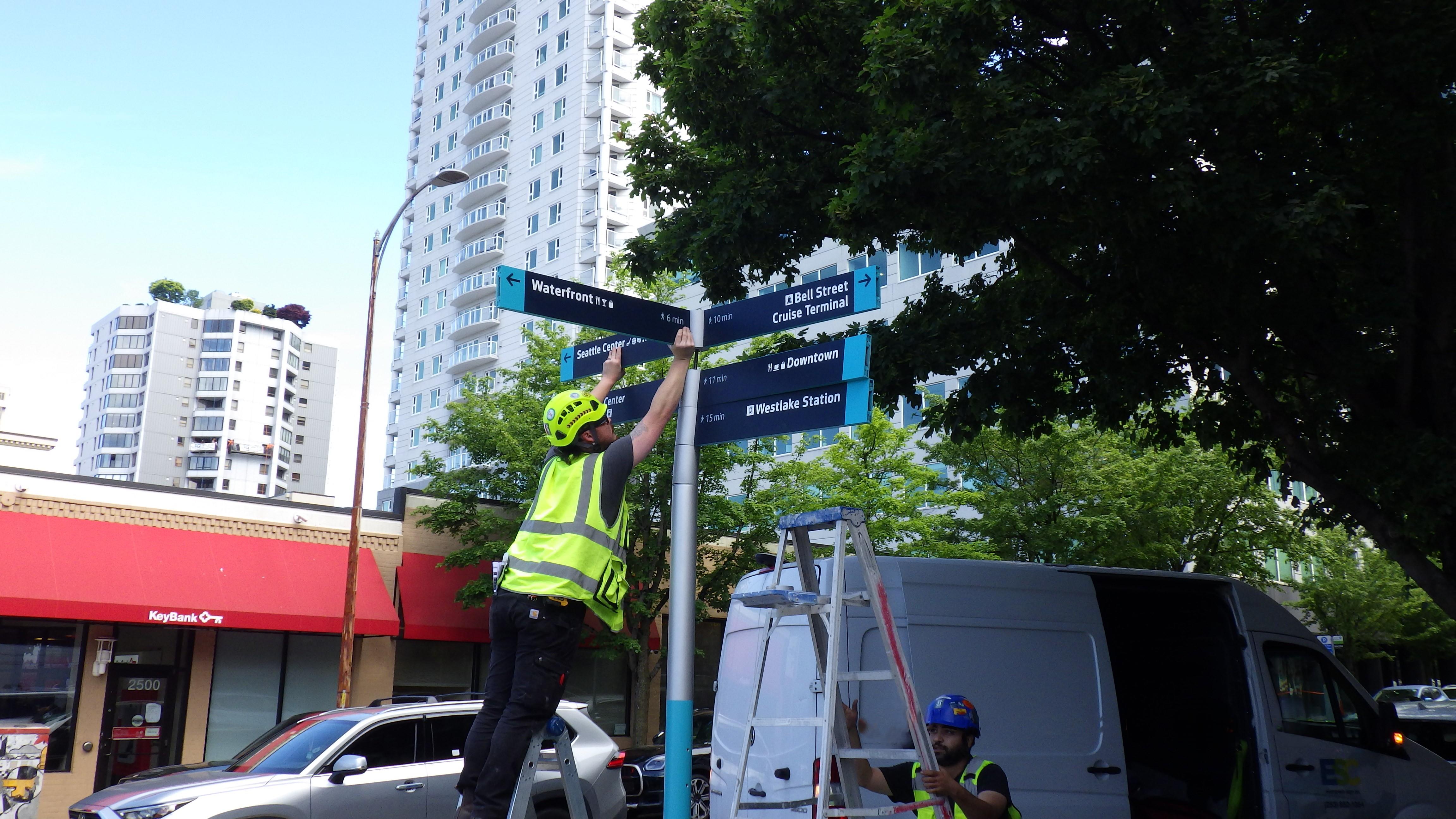 A construction worker in a yellow vest and helmet installs a wayfinding sign on a pole in downtown Seattle. The sign displays directions to nearby destinations including the Waterfront, Bell Street Cruise Terminal, Seattle Center, Downtown, and Westlake Station. A ladder and work van are visible in the background. 