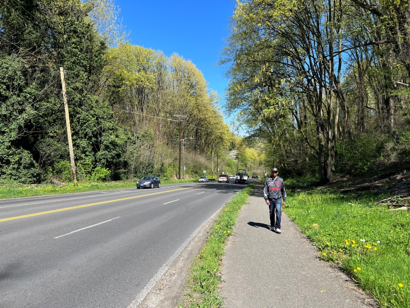 A person walking on the existing path along Highland Park Way SW. To the left of the path are two with two northbound driving lanes and two southbound driving lanes. There are cars driving in both directions. 