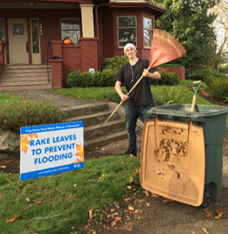 A man holds a rake in a yard with a yard waste bin and a yard sign reading RAKE LEAVES TO PREVENT FLOODING.