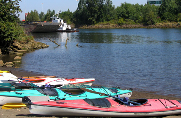 A sunny day with kayaks beached on the banks of the Duwamish with commercial vessels in background.