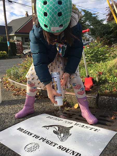 A child wearing a helmet and rubber boots standsA young girl wearing a green bike helmet and pink boots spray paints a stenciled message near a street drain.