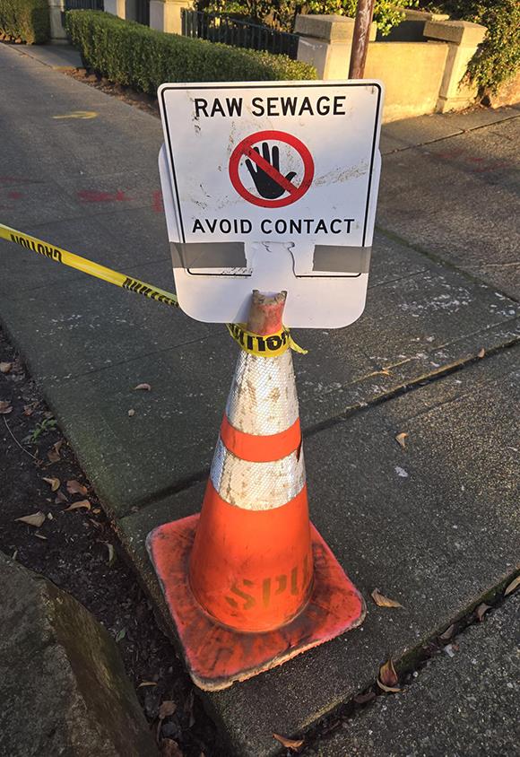 An orange traffic cone with yellow caution tape tied to it on a street corner with a sign on top of it that reads, "RAW SEWAGE. AVOID CONTACT."