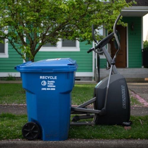 An exercise machine on a parking strip near a recycling bin.