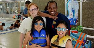 Four individuals standing beside an indoor pool, with two wearing life jackets and goggles.