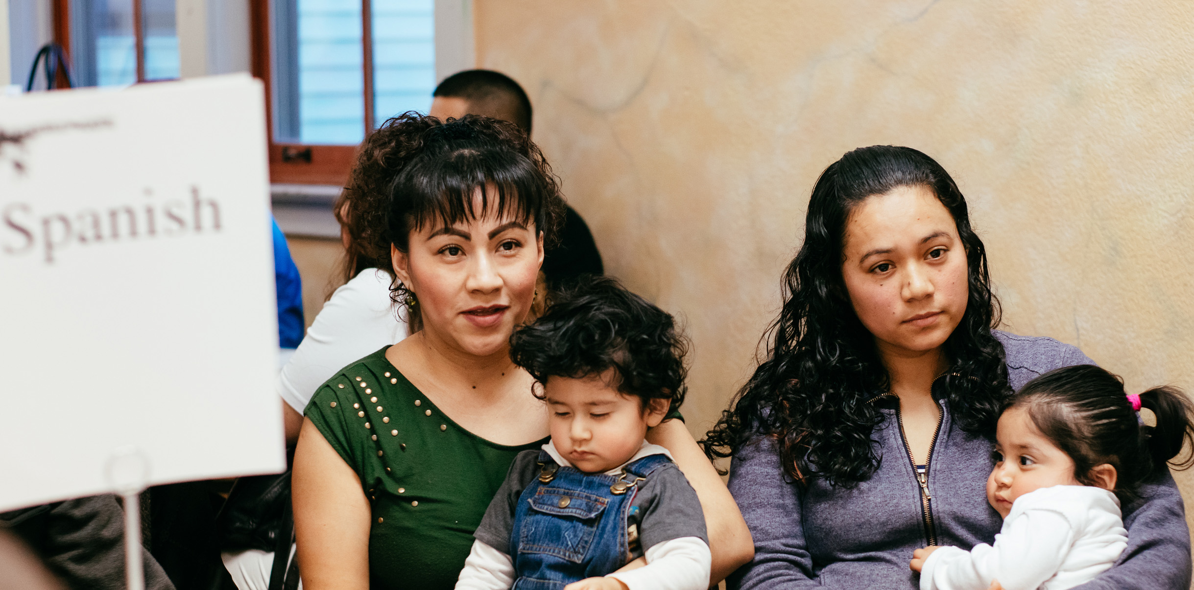 Two Seattle Latina women, each holding a baby, appear to be talking to someone next to a sign that reads: "Spanish".