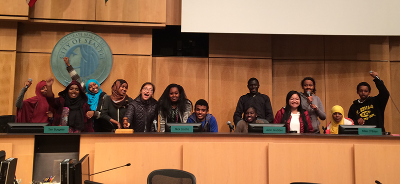 A group of multiracial young people sitting in the councilmember seats in Seattle City Council Chambers. Some youths are in humorous poses.