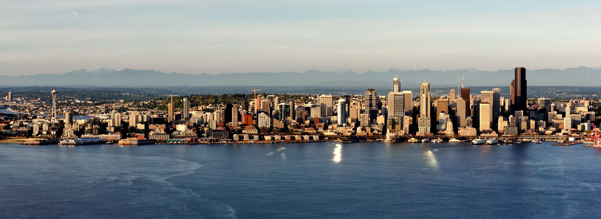 Wide panoramic view of a waterfront city skyline with tall buildings, a prominent tower, and mountains in the background across a calm body of water.