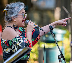 Profile shot of a singer pointing with their left hand. She's wearing shades, an off-the-shoulder colorful top, and dangly earrings.