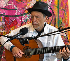 An older Latino wearing a black hat and white long-sleeved shirt plays the guitar against a colorful painted background.