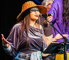 A singer with long hair and a traditional woven hat stands at the mic with palms open upwards. She's wearing silver bangles and a fabric belt with a Native carving pattern.