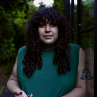 Jesse has long, dark, curly hair, bangs, and tattoos. They're smizing in a forest background.