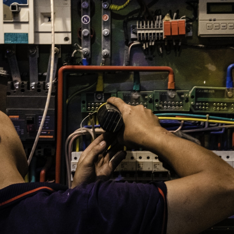 A person is working on an electrical panel, connecting wires. The individual’s focus is on the task at hand, with their hands manipulating cables within the open panel which contains various electrical components.