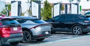 Two electric vehicles, one red SUV and one silver sedan, parked at white EV charging stations with charging cables connected, with a background of trees and urban buildings