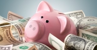 A pink piggy bank is surrounded by various denominations of US dollar bills against a light blue background. The image represents savings, highlighting the traditional method of saving money in a piggy bank.