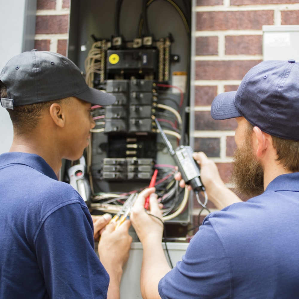 Two individuals are working on an open electrical panel attached to a brick wall. One individual is holding a multimeter to the wires inside the panel, while the other is observing. The faces of both individuals are not visible in the image. The focus is on their hands and the equipment they are using, indicating they are conducting an electrical inspection or repair.