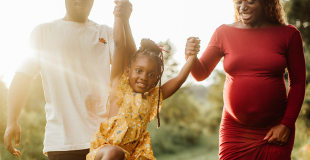 An expecting Black parent, wearing a red dress, and her partner, wearing a white t-shirt, hold hands with their preschool age child, wearing a yellow sundress