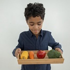 Young boy holding a tray with vibrantly colored fresh foods, including an ear of yellow corn, orange carrot, red apple, and green broccoli stalk
