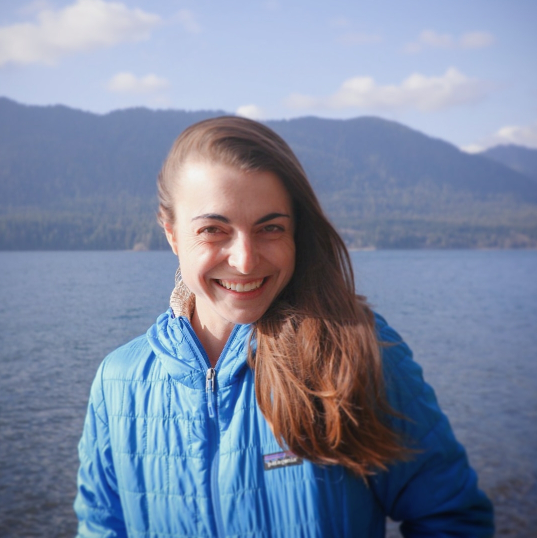 Close up of Nina Olivier from the elbows up smiling in coat with mountains in the background.  