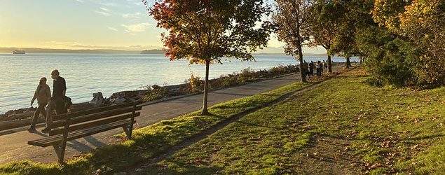 People walking on a paved path along the waterfront; sun is starting to set