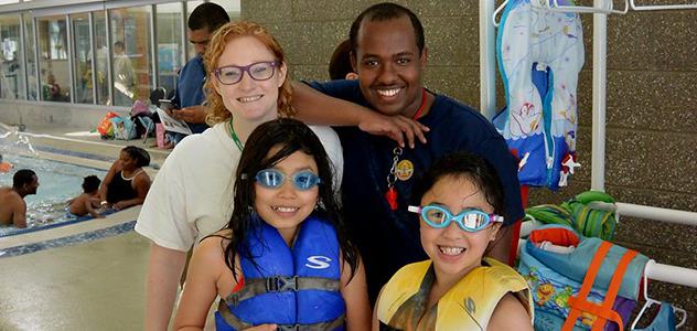 Four individuals standing beside an indoor pool, with two wearing life jackets and goggles.