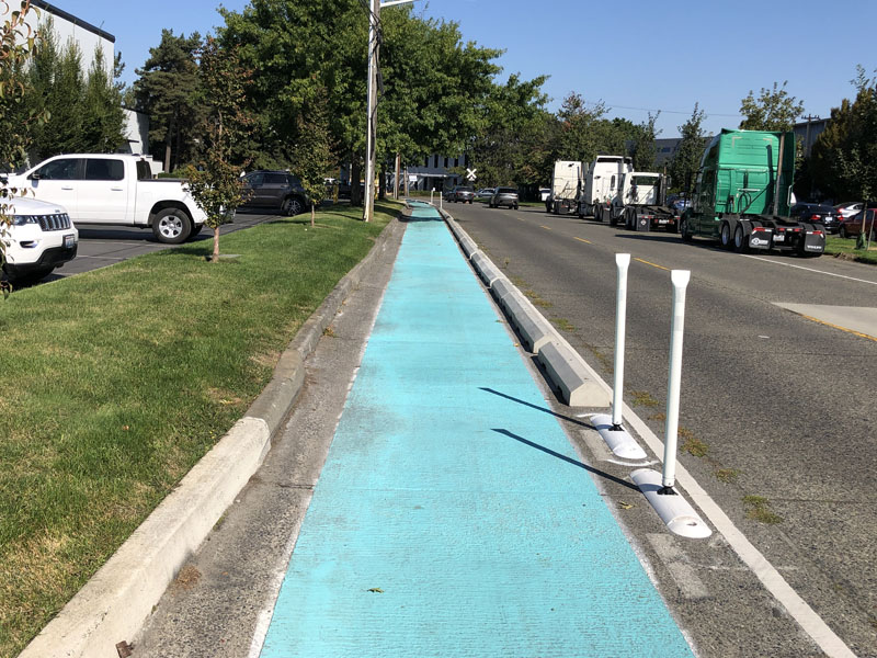 A bright blue painted sidewalk with wheel stop delineators and flex-posts