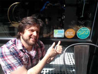 Photo of owner of Delicatus restaurant giving thumbs in front of the window with green business stickers.