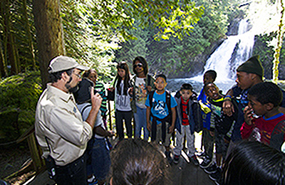 An instructor speaks to a group of students in a wooded area next to Cedar Falls.