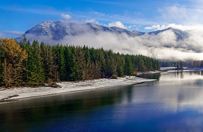 Evergreen forest lines the rocky shores of the Cedar River with mist covered mountains in the background under a clear blue sky.