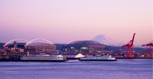 A view of the Seattle waterfront at dusk, featuring two large ferries, cargo cranes, and a view of Lumen Field and Mt. Ranier against a pink and purple sky.