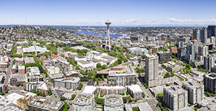 Panoramic aerial view of the Seattle cityscape on a clear day, showcasing a dense arrangement of various high-rise buildings. To the left, the Space Needle stands out against the blue sky, while to the right, the urban architecture transitions into a waterfront with numerous boats and docks visible. The natural landscape includes vast water bodies and greenery surrounding the metropolitan area.