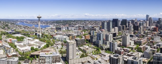 Panoramic aerial view of the Seattle cityscape on a clear day, showcasing a dense arrangement of various high-rise buildings. To the left, the Space Needle stands out against the blue sky, while to the right, the urban architecture transitions into a waterfront with numerous boats and docks visible. The natural landscape includes vast water bodies and greenery surrounding the metropolitan area.