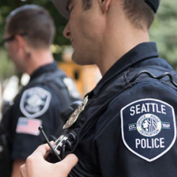 Close-up of a Seattle Police Department uniformed police officer holding a shoulder-mounted radio, with a visible patch that reads ‘Seattle Police.’ Another officer is slightly out of focus in the background outdoors.